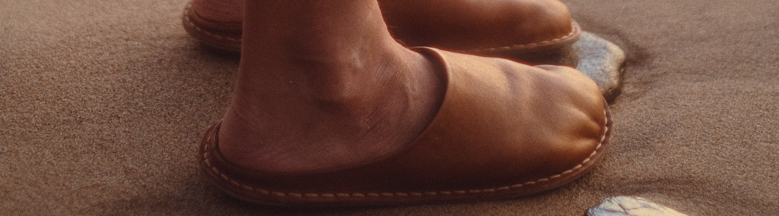 Ett par fötter i brunbeige barfotaskor av modellen VIBAe Roma, stående på sand med synliga stenar. Skorna har rundad tåform och synlig söm längs sulkanten.