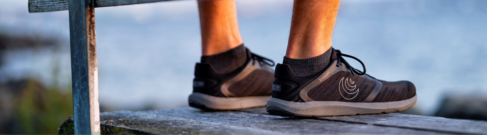 Ett par svarta löparskor med grå sula från Topo, modellen ST-5. Skorna bärs av en person stående på en träbrygga vid vatten, med kvällsljus och suddig bakgrund. Tydlig logotyp syns på sidan av skon.