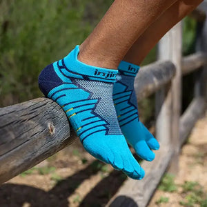 Person med blå Injinji tåstrumpor sittandes på ett trästaket i naturmiljö. Strumporna har individuell tåpassform och färgstarkt mönster för optimal komfort vid löpning och vandring