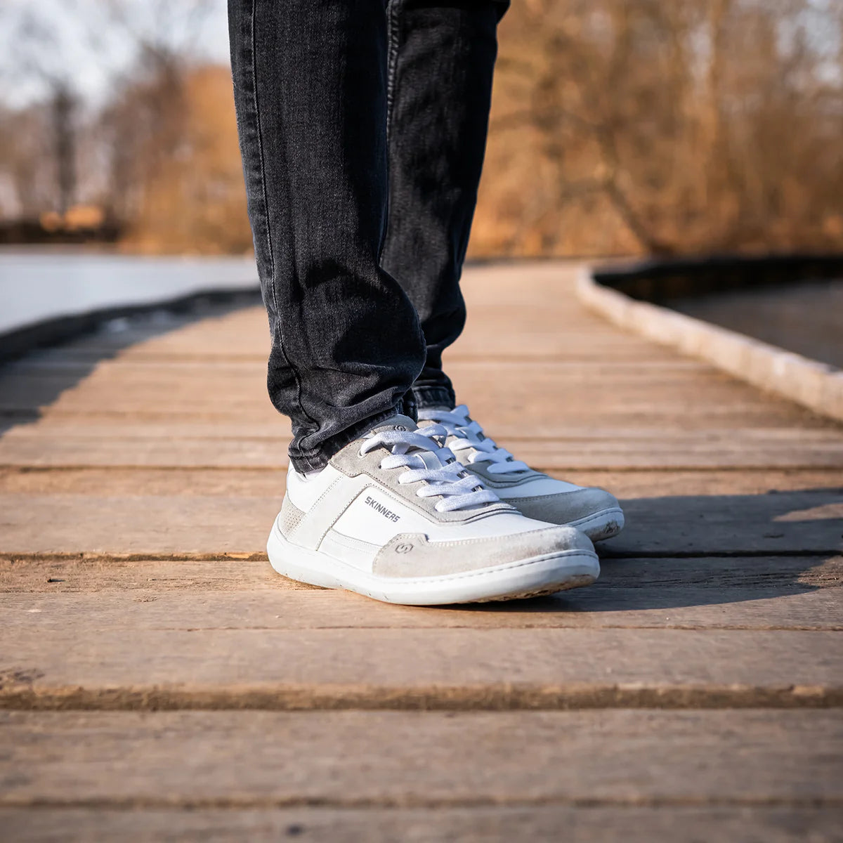 Person står på en brygga iförd Skinners Allrounder White barfotaskor – minimalistiska sneakers i vitt och ljusgrått med bred tåbox, perfekt för naturlig rörelse året runt.