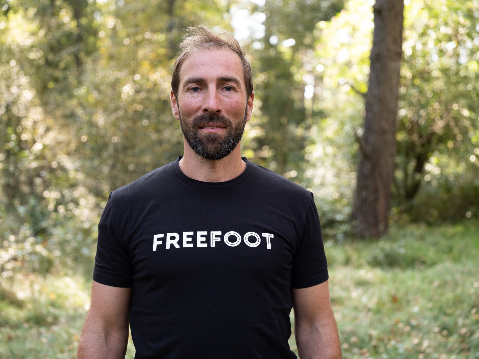 Man i svart Freefoot-t-shirt står i en solbelyst skogsmiljö, tittar in i kameran. Profilbild för varumärket Freefoot.