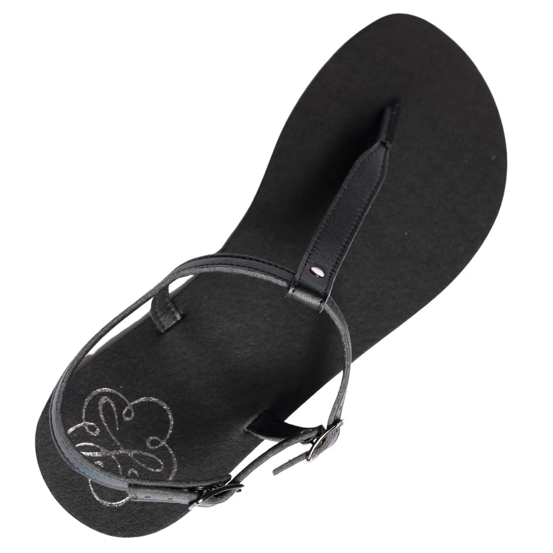 Produktbild på Lonnies barfotasko i modellen Lux Little Black, fotograferad i sned vinkel framifrån. Sandalen har tunna svarta remmar med spänne, T-remdesign och en minimalistisk svart sula. Perfekt för barfotakänsla med stilren look