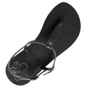 Produktbild på Lonnies sandaler Lux Little Black sedd uppifrån. Den minimalistiska barfotaskon har svarta remmar i T-design, ett justerbart spänne och en slät svart sula med Lonnies logotyp tryckt i hälen. Designad för barfotakänsla med stilren komfort.