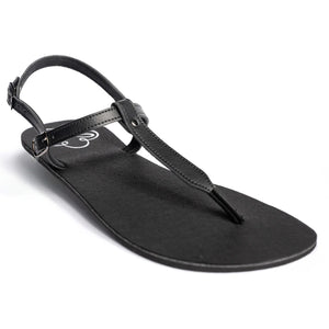 Produktbild på Lonnies barfotasko i modellen Lux Little Black, fotograferad i sned vinkel framifrån. Sandalen har tunna svarta remmar med spänne, T-remdesign och en minimalistisk svart sula. Perfekt för barfotakänsla med stilren look