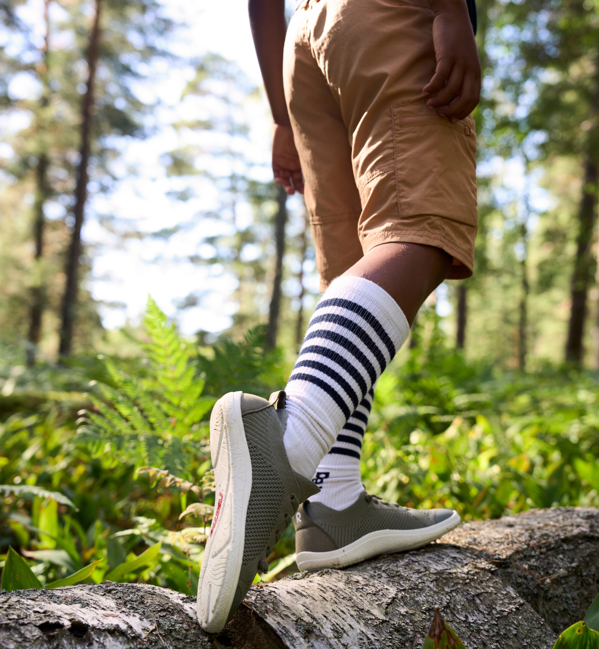Ett barn går på en stock i skogen iförd barfotaskor från Reima. Skorna har en bred tåbox och flexibel sula som uppmuntrar till naturlig rörelse i utomhusmiljö.