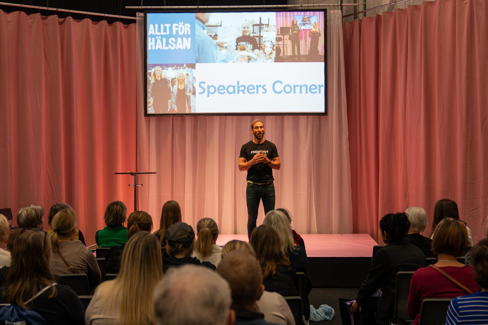 Föreläsare i svart Freefoot-t-shirt står på scen under mässan Allt för Hälsan, inför sittande publik vid Speakers Corner.